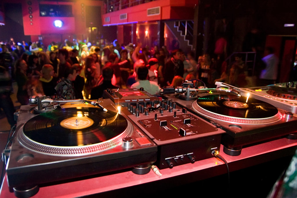A DJ booth with turntables in front of a crowded dance floor at a club.