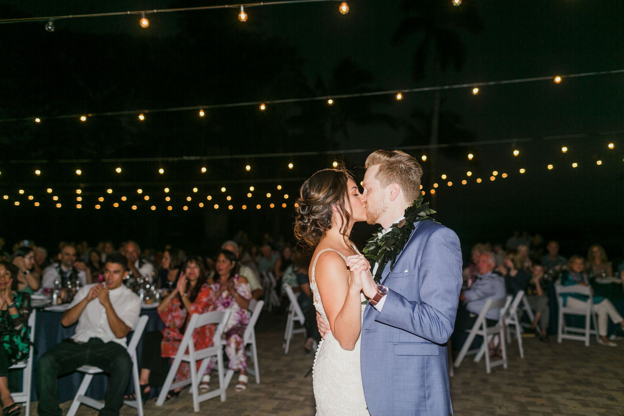 Bride and groom kiss during their first dance under string lights at night.